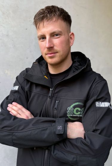 Portrait von Nick Heyden, Meister im Gebäudereiniger-Handwerk, mit Arbeitsjacke und SCS Logo
